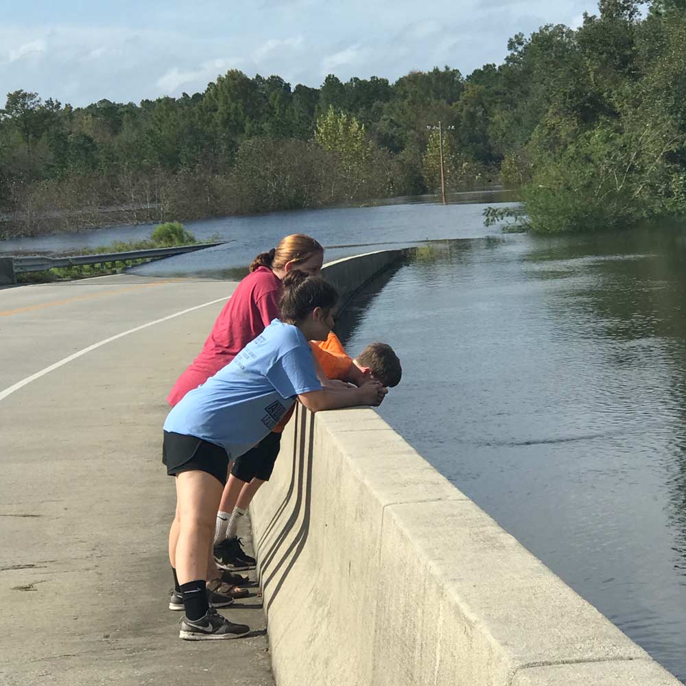 Tres niños se paran al borde de una carretera inundada mirando hacia el agua.