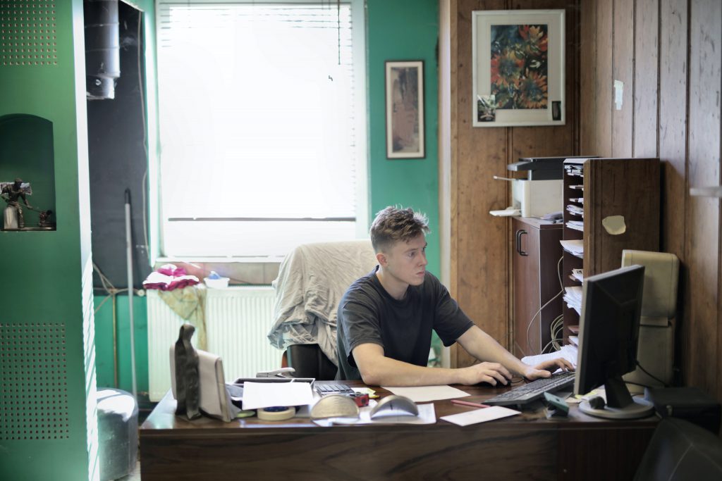A man in a T-shirt works on a computer at a cluttered desk/